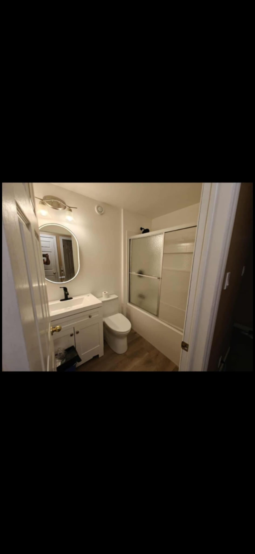 Compact bathroom with round backlit mirror, white vanity, and sliding tub door