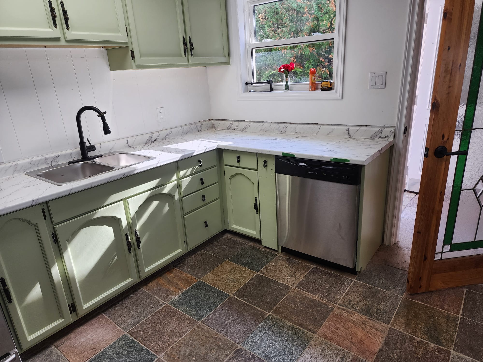 Sage green kitchen cabinetry with marble-look countertop, black faucet, and dishwasher by the window